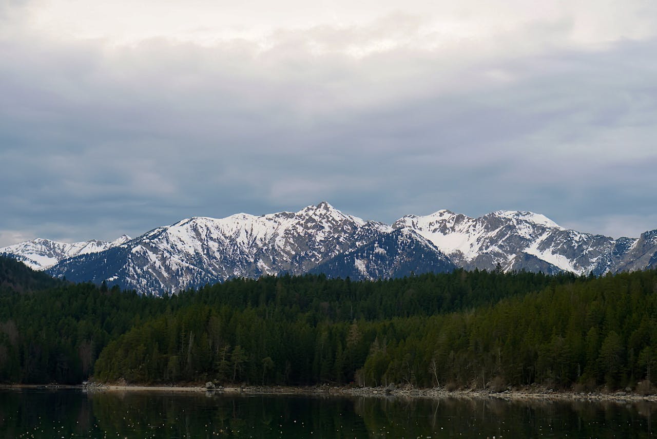 Breathtaking view of snowcapped mountains above pine forests reflected in calm waters.